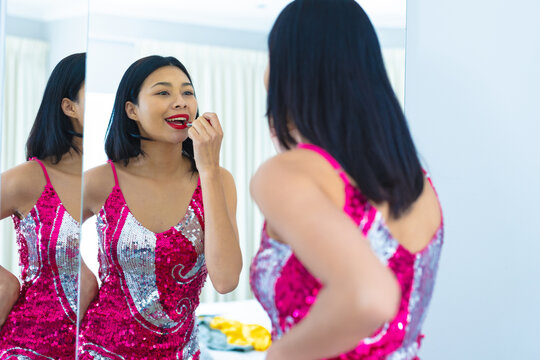 Happy biracial woman in party dress looking in mirror, putting on lipstick, copy space