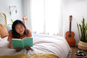 Smiling Asian young woman reading book lying on bed, relaxing in cozy bedroom looking at camera.