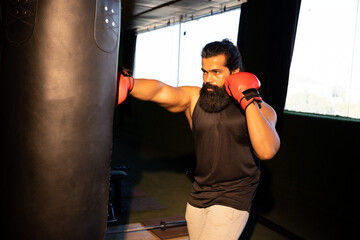 Young indian beard man wearing boxing gloves and hooded sweatshirt punching bag in gym, Fitness and healthcare.