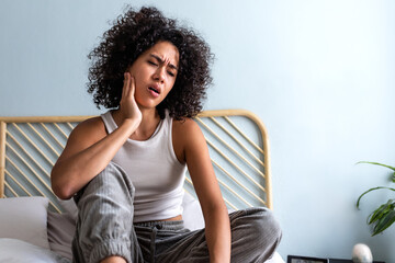 Young latina female with curly hair touching cheek in pain having toothache sitting on bed in cozy...