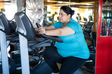Young overweight indian woman is workout in gym to lose weight. Asian female doing cardio training on modern bike bicycle machine.