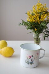 Enameled painted pot, lemon and yellow mimosa flowers in a glass vase on white table