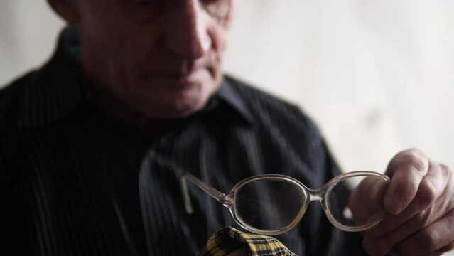 an elderly man wipes his glasses from dirt and dust with a napkin. a pensioner cleans the lenses of glasses. loss of vision in old age. selective focus. lifestyle of an old man. eye fatigue