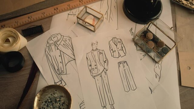 Cinematic shot of table with drawings, needles, threads, buttons, ribbons, soap and ruler. Sketches of clothes, suits and jackets. Designer atelier or tailoring studio. Fashion and hand craft concept.