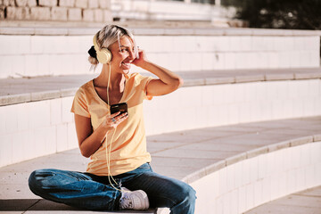 Woman looking away and smiling while listening music with headphones sitting outdoors.