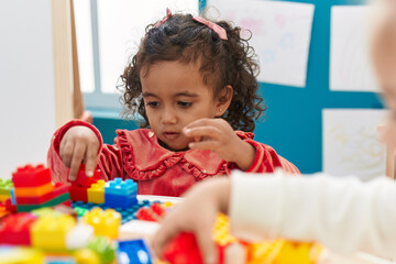 Adorable hispanic girl playing with construction blocks sitting on table at kindergarten