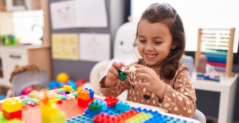 Adorable hispanic girl playing with construction blocks sitting on table at kindergarten