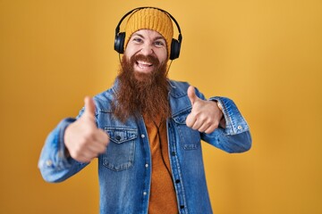 Caucasian man with long beard listening to music using headphones approving doing positive gesture with hand, thumbs up smiling and happy for success. winner gesture.