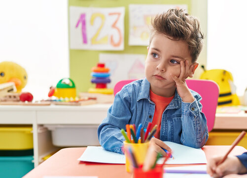 Adorable hispanic toddler student sitting on table drawing on paper with doubt expression at kindergarten