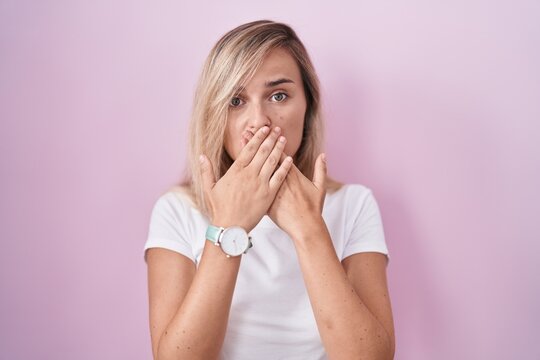 Young blonde woman standing over pink background shocked covering mouth with hands for mistake. secret concept.
