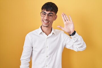 Young hispanic man standing over yellow background waiving saying hello happy and smiling, friendly welcome gesture