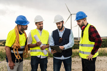 Team of engineers and technical workers working outdoors wind turbine farm planning construction project with blueprint map and tablet
