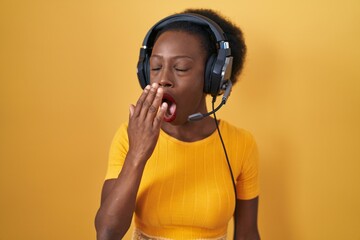 African woman with curly hair standing over yellow background wearing headphones bored yawning tired covering mouth with hand. restless and sleepiness.