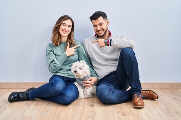 Obraz na płótnie Canvas Young hispanic couple sitting on the floor with dog cheerful with a smile on face pointing with hand and finger up to the side with happy and natural expression