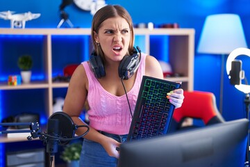 Young blonde woman playing video games holding keyboard angry and mad screaming frustrated and...