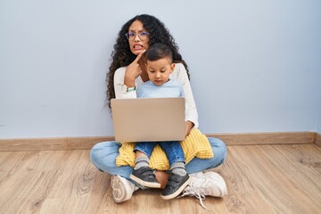 Young hispanic mother and kid using computer laptop sitting on the floor pointing aside worried and...