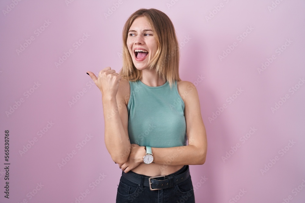 Poster blonde caucasian woman standing over pink background smiling with happy face looking and pointing to