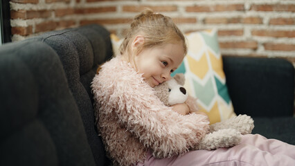 Adorable blonde girl hugging teddy bear sitting on sofa at home