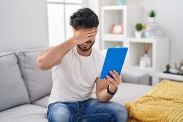 Fototapeta premium Hispanic man with beard using touchpad sitting on the sofa smiling and laughing with hand on face covering eyes for surprise. blind concept.