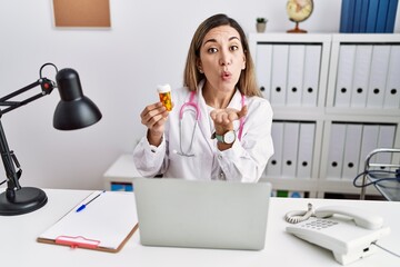 Young hispanic woman wearing doctor uniform holding pills at the clinic looking at the camera blowing a kiss with hand on air being lovely and sexy. love expression.