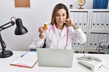 Young hispanic woman wearing doctor uniform holding pills at the clinic mouth and lips shut as zip with fingers. secret and silent, taboo talking