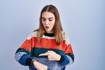 Young hispanic girl standing over blue background in hurry pointing to watch time, impatience, upset and angry for deadline delay