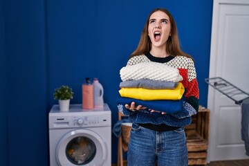 Young hispanic girl holding clean laundry angry and mad screaming frustrated and furious, shouting with anger looking up.