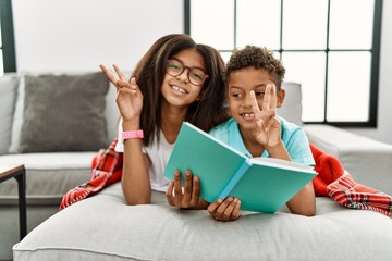 Two siblings lying on the sofa reading a book smiling looking to the camera showing fingers doing...