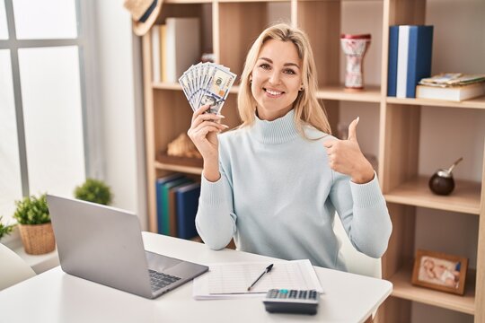 Young caucasian woman using laptop holding dollars banknotes smiling happy and positive, thumb up doing excellent and approval sign