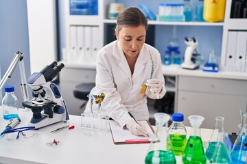 Middle age woman scientist holding test tube with plant writing on clipboard at laboratory