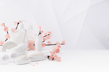 Cosmetic and beauty products for bath, spa in white bottles, branch of spring pink sakura flowers, toiletry on table in soft light white bathroom interior in geometric simple urban style, copy space.