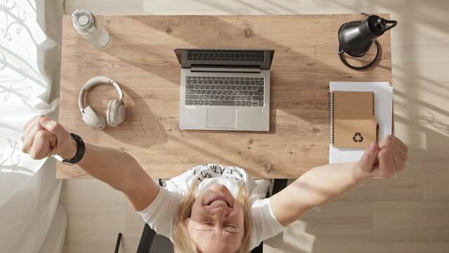 Top down view of girl working at computer, achieving success and joy. young woman throws her hands up in win or successful deal. blonde in white t-shirt rejoices while sitting at workplace.