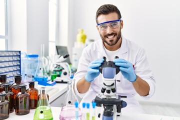 Young hispanic man wearing scientist uniform using microscope at laboratory