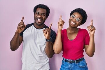 Young african american couple standing over pink background smiling amazed and surprised and pointing up with fingers and raised arms.