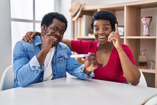 Young african american couple holding pregnancy test result surprised with an idea or question pointing finger with happy face, number one