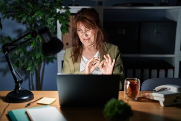 Middle age hispanic woman working using computer laptop at night disgusted expression, displeased and fearful doing disgust face because aversion reaction. with hands raised