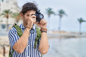 Young hispanic man tourist wearing backpack using professional camera at seaside