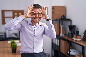 Young hispanic man at the office doing ok gesture like binoculars sticking tongue out, eyes looking through fingers. crazy expression.