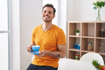 Young hispanic man drinking coffee sitting on table at home