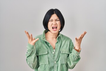 Young asian woman standing over white background crazy and mad shouting and yelling with aggressive expression and arms raised. frustration concept.