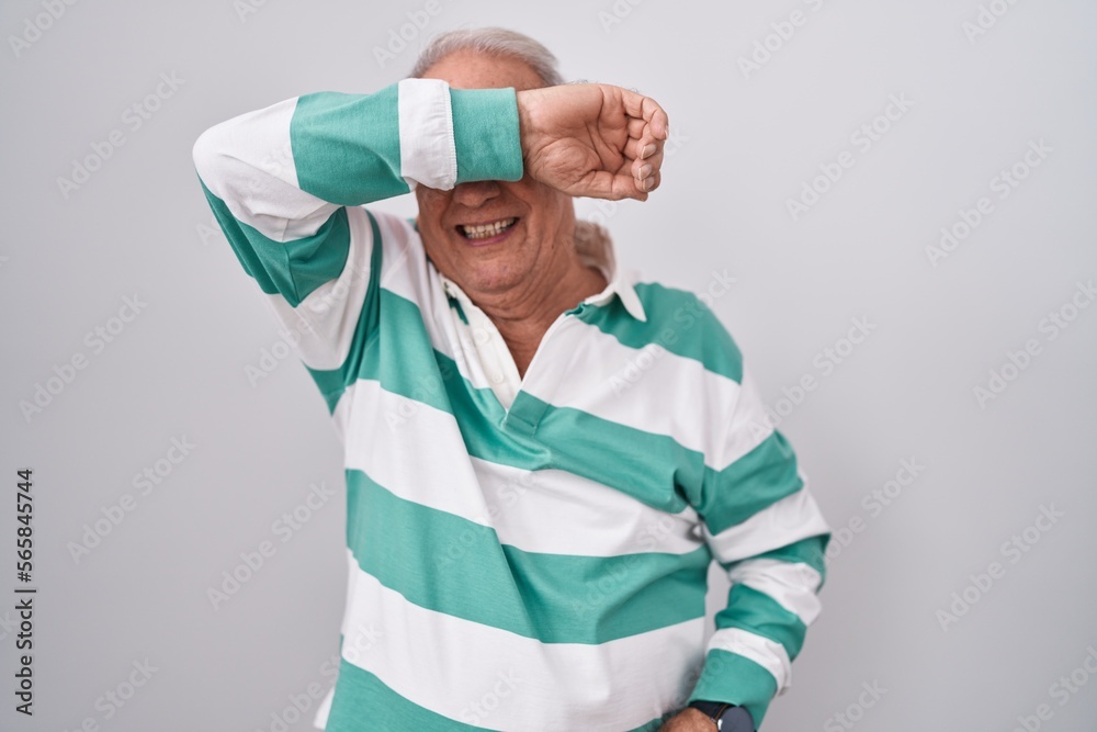 Poster senior man with grey hair standing over white background covering eyes with arm smiling cheerful and