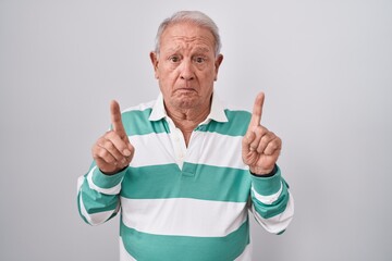 Senior man with grey hair standing over white background pointing up looking sad and upset, indicating direction with fingers, unhappy and depressed.