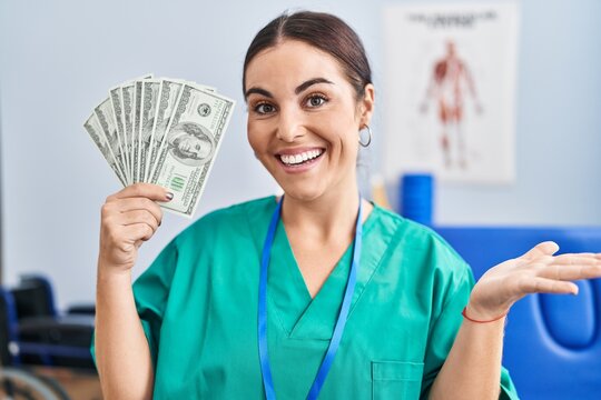 Young hispanic woman holding dollars banknotes working at pain recovery clinic celebrating achievement with happy smile and winner expression with raised hand