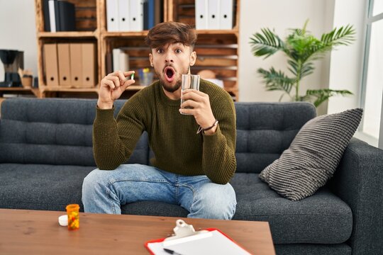 Arab man with beard working on depression holding pills and water afraid and shocked with surprise and amazed expression, fear and excited face.