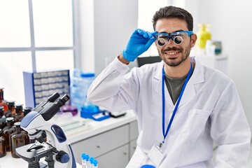 Young hispanic man working at scientist laboratory wearing magnifying glasses looking positive and happy standing and smiling with a confident smile showing teeth