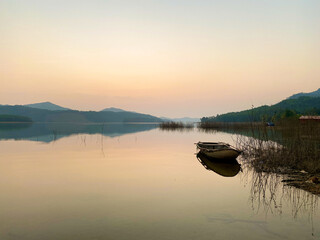 Peaceful summer sunset with calm lake surface reflecting green mountains and an anchored boat. Tranquil natural background