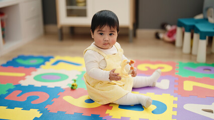 Adorable hispanic baby holing toy sitting on floor at kindergarten