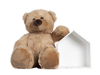 Cute new light brown toy teddy bear sitting facing front beside little wooden house. Isolated cutout on transparent background.