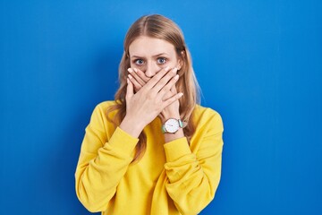 Young caucasian woman standing over blue background shocked covering mouth with hands for mistake. secret concept.