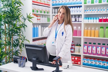 Young pregnant woman pharmacist talking on smartphone using computer at pharmacy